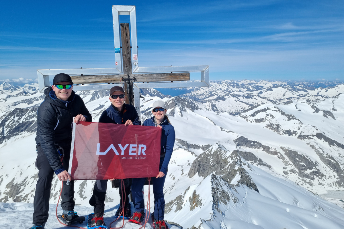 Zwei M&auml;nner und eine Frau mit Sonnenbrillen am Gipfelkreuz im Schnee mit roter Layer-Fahne