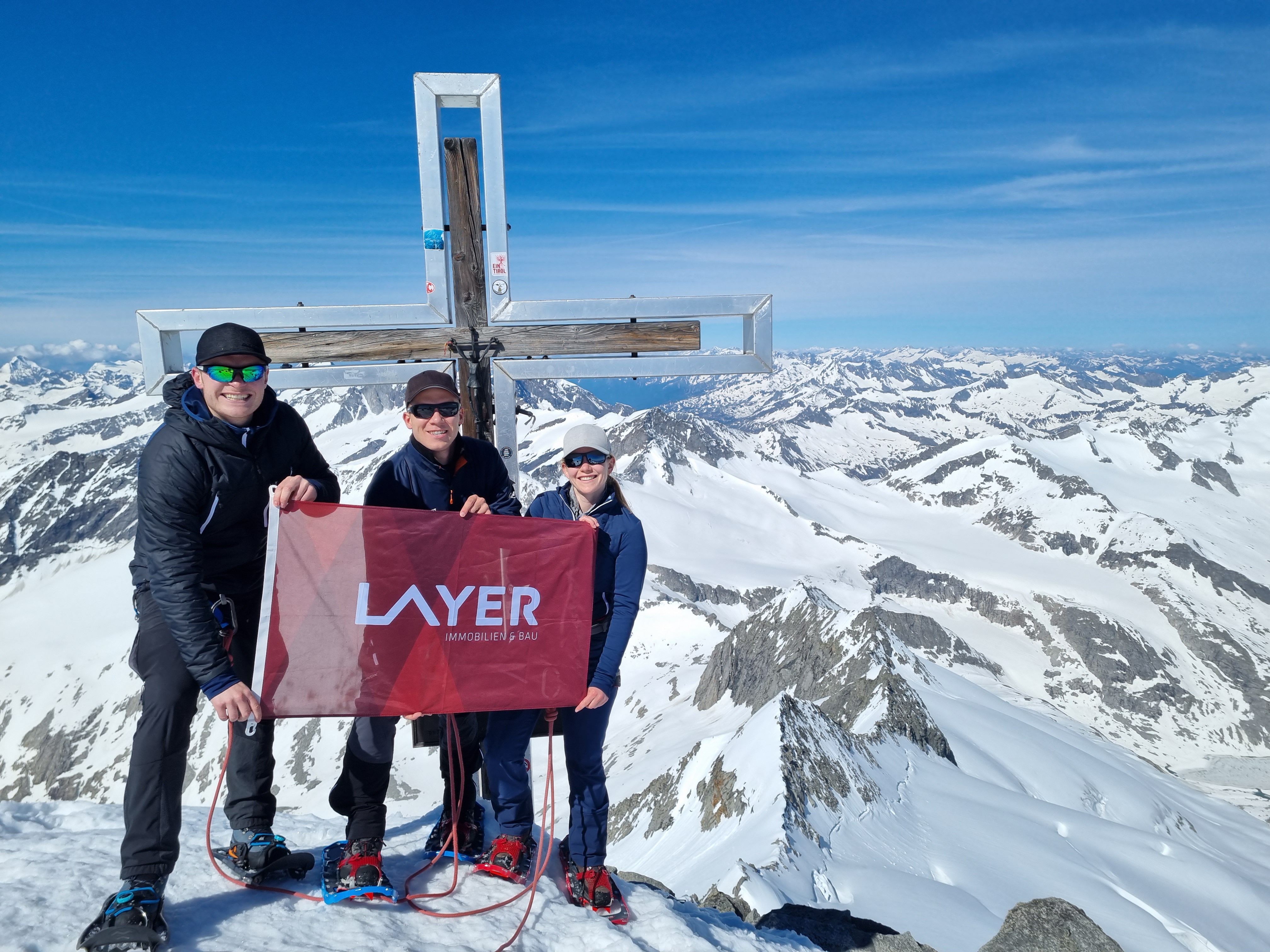 Zwei M&auml;nner und eine Frau mit Sonnenbrillen am Gipfelkreuz im Schnee mit roter Layer-Fahne