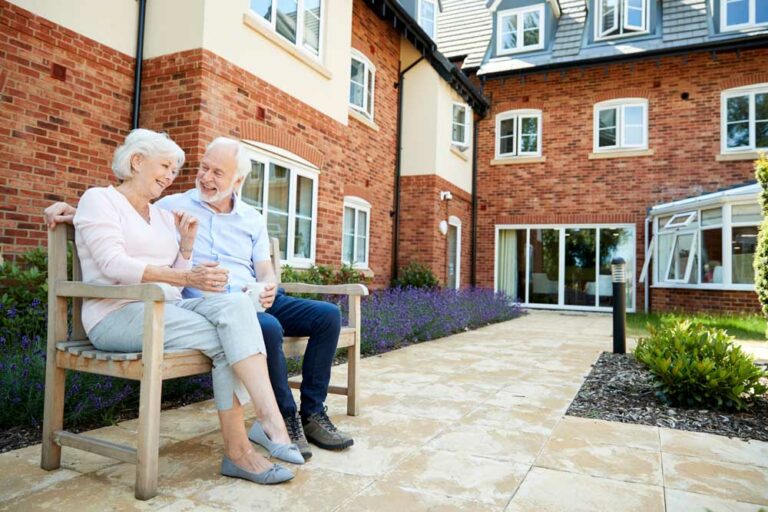 Ein lachendes Ehepaar, dass sich für eine Pflegeimmobilie als Kapitalanlage entschieden hat, sitzt im eigenen Ruhestand auf einer Bank vor einer Wohnanlage und führt eine Unterhaltung
